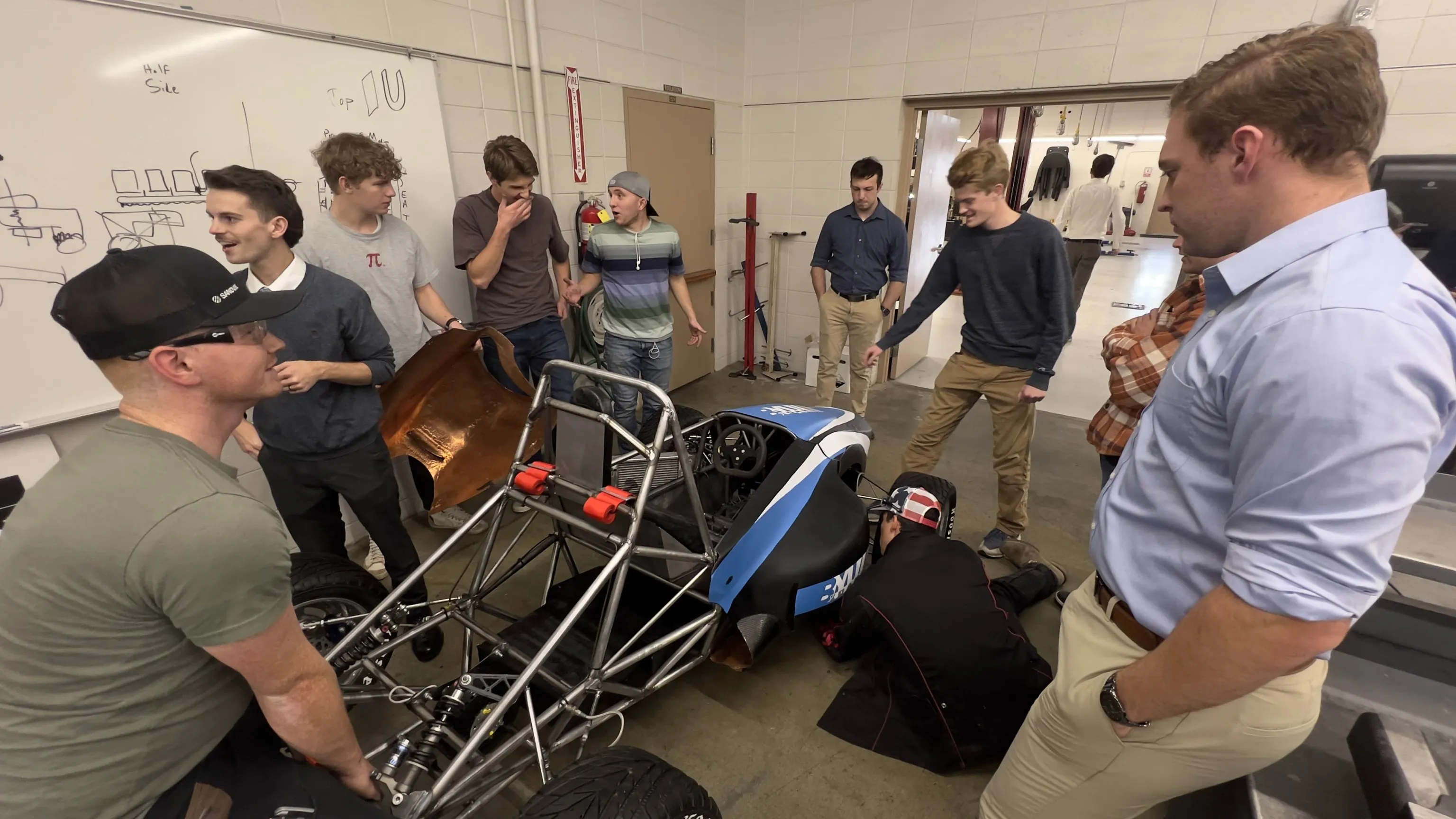 A group gathered around a blue and white formula car with an exposed chassis in the workshop